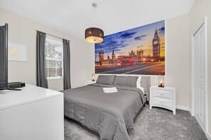 A bedroom with a large bed, a desk, a nightstand, a lamp, and a painting of a cityscape.