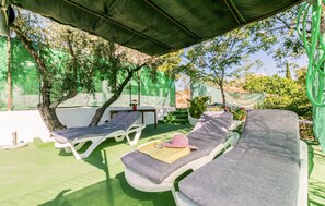 A covered outdoor seating area with two lounge chairs and a table, surrounded by greenery and a fence.