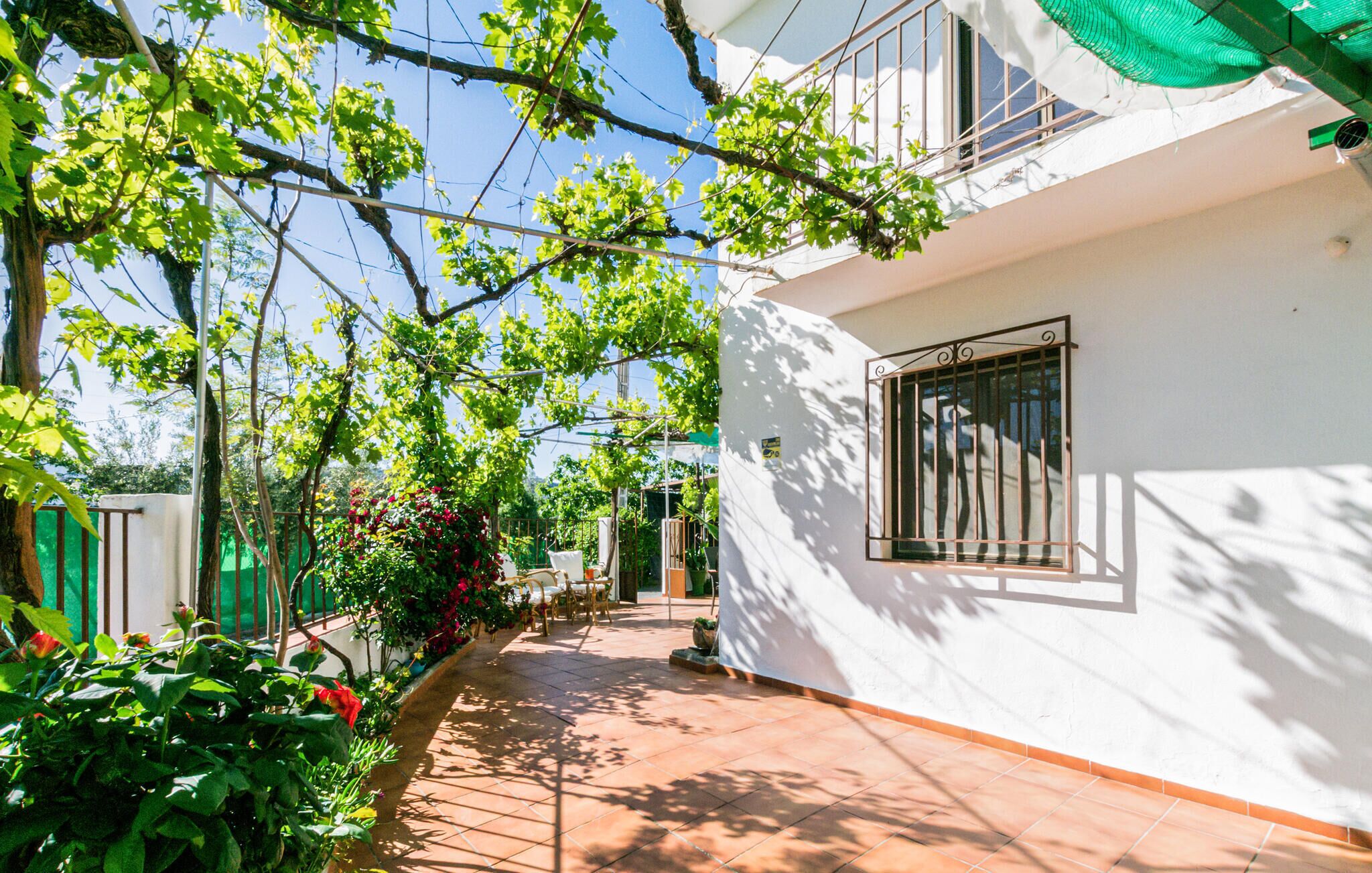 A covered outdoor area with a tiled floor, surrounded by greenery and a white building with a window.