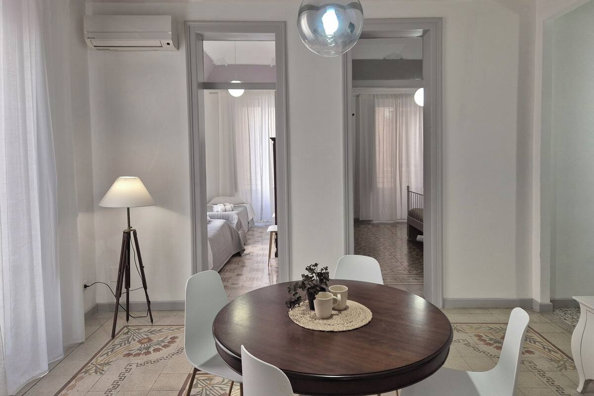 A hotel room with a round dining table, white chairs, a lamp, and a bed with a canopy.