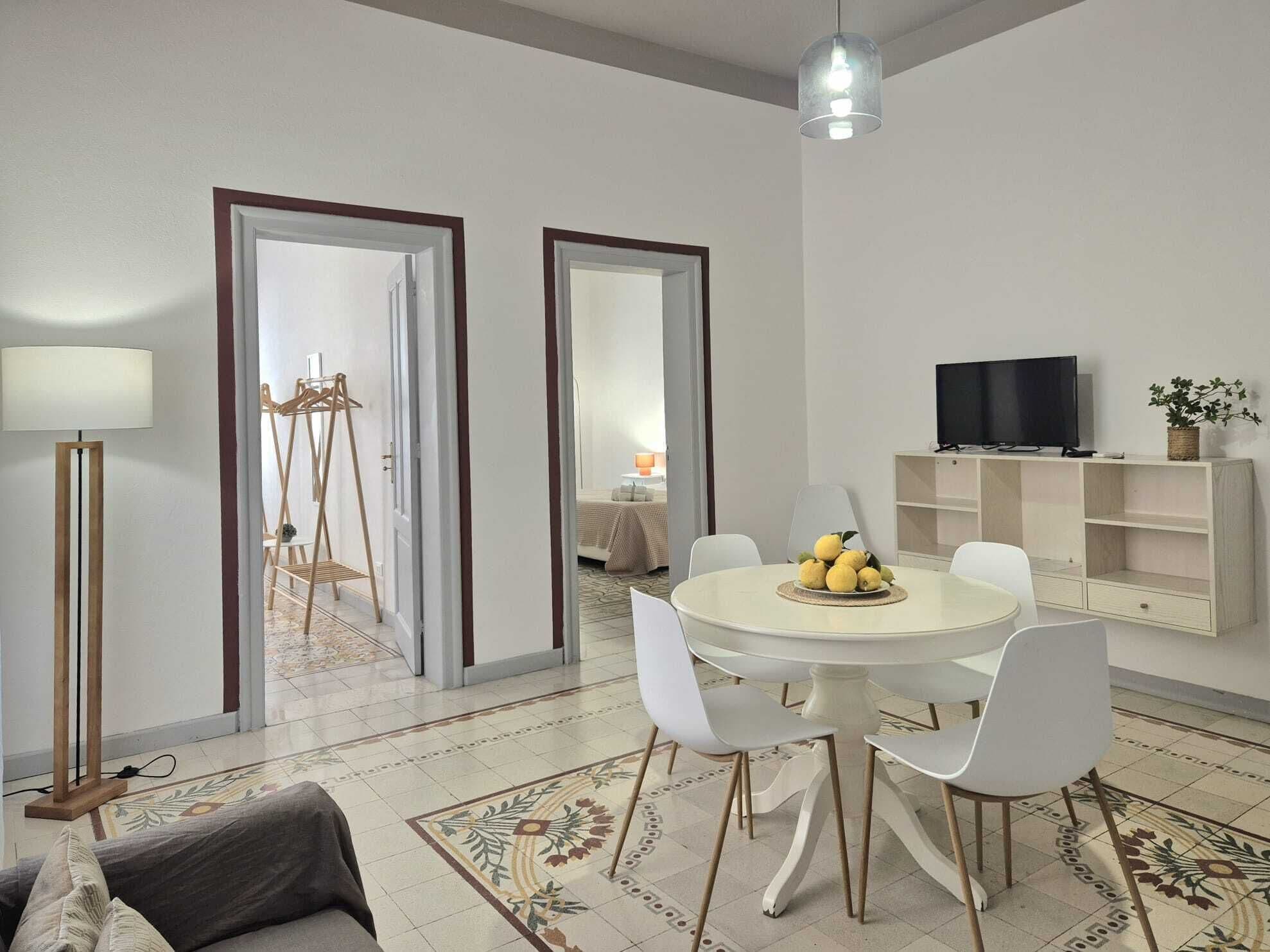A modern living room with a round dining table, white chairs, a TV on a shelf, and a floor lamp.