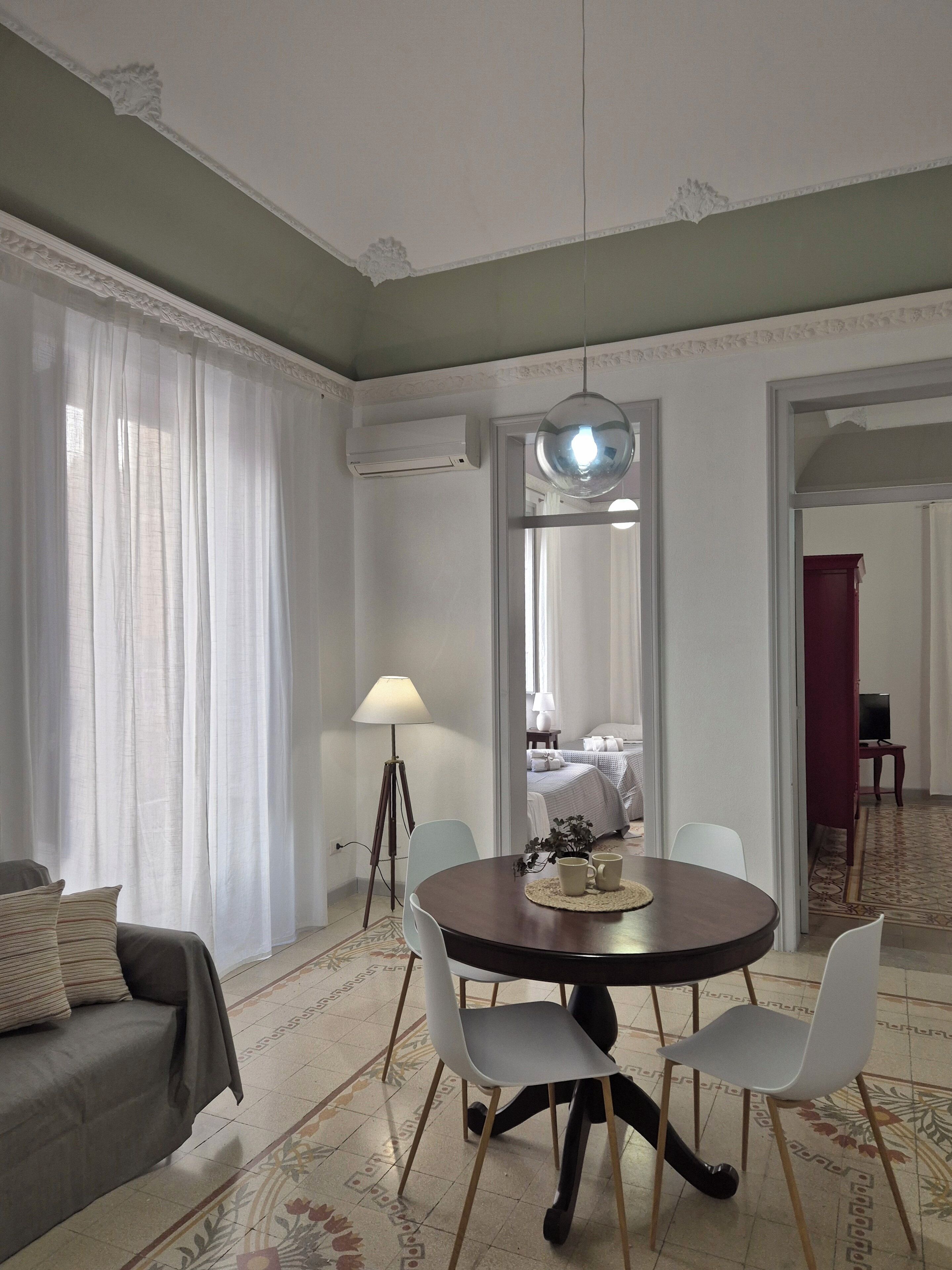 Ein modernes Wohnzimmer mit einem runden Esstisch, zwei weißen Stühlen, einem grauen Sofa, einer Stehlampe und einem großen Spiegel.