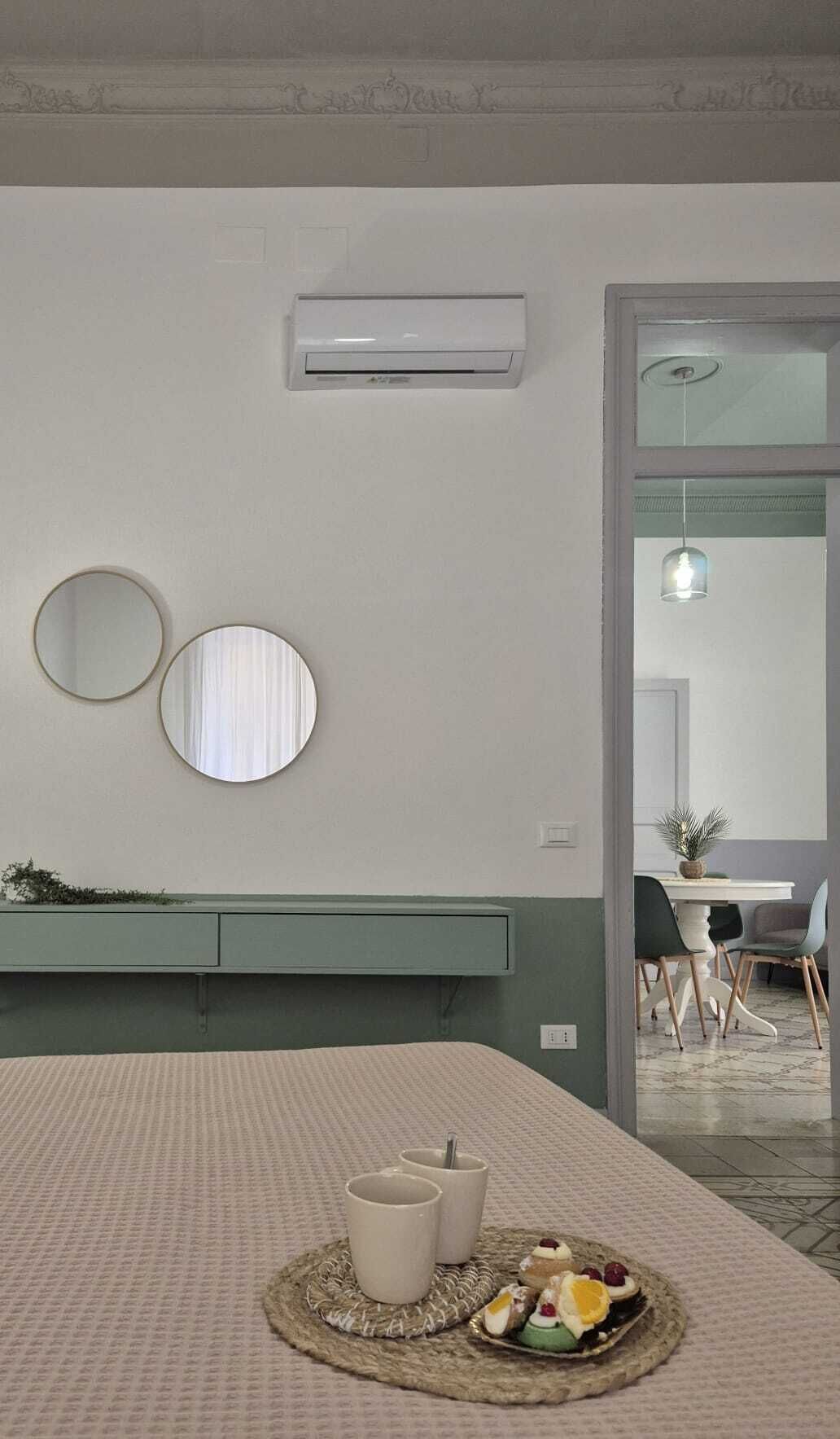 Una stanza con un letto, un comodino con uno specchio, un condizionatore d'aria a parete e un'area pranzo visibile attraverso una porta aperta.