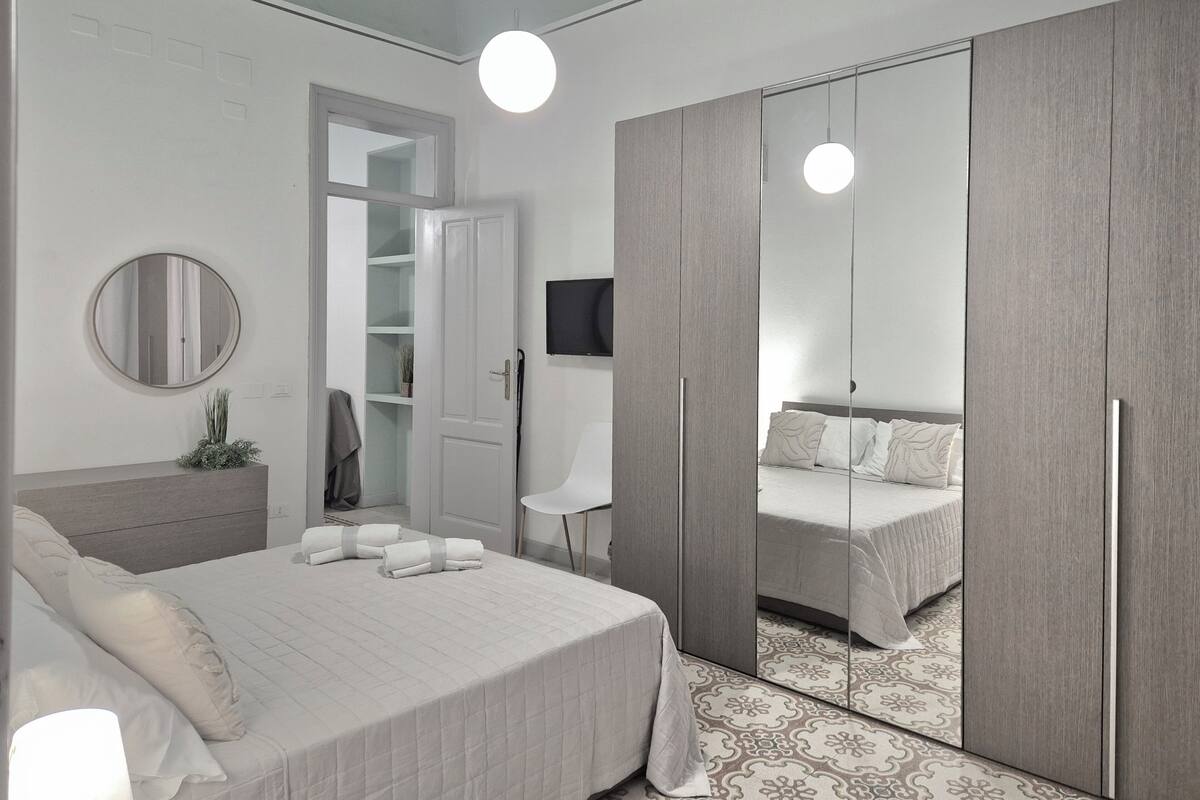 A modern bedroom with a bed, bedside table, mirror, wardrobe, and a TV.