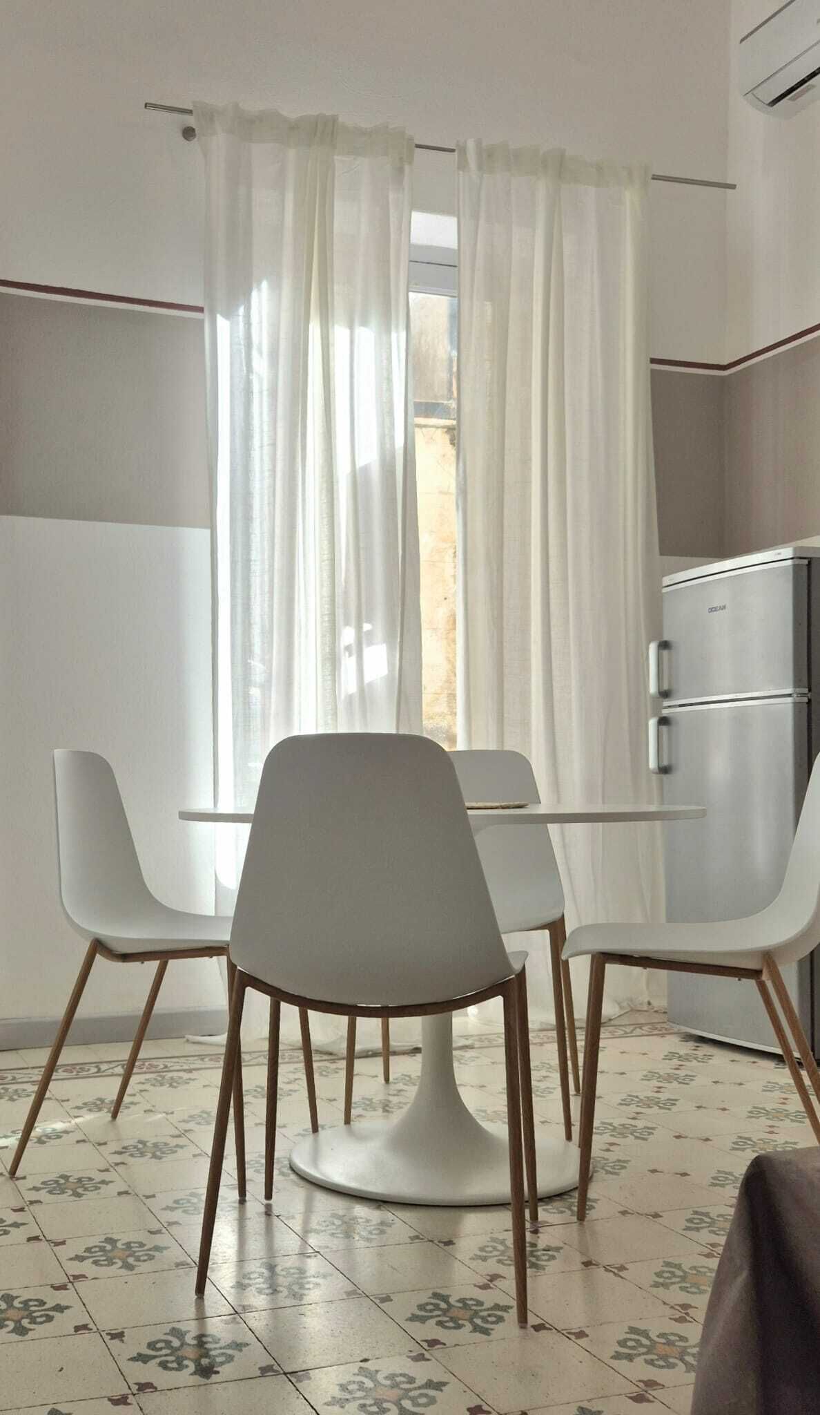 A modern room with a dining table and chairs, a kitchen area, and a window with sheer curtains.
