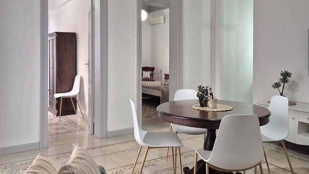 A modern living room with a round dining table, white chairs, a sofa with striped cushions, and a patterned rug. - CASE VACANZE LA MATTANZA (Trapani)