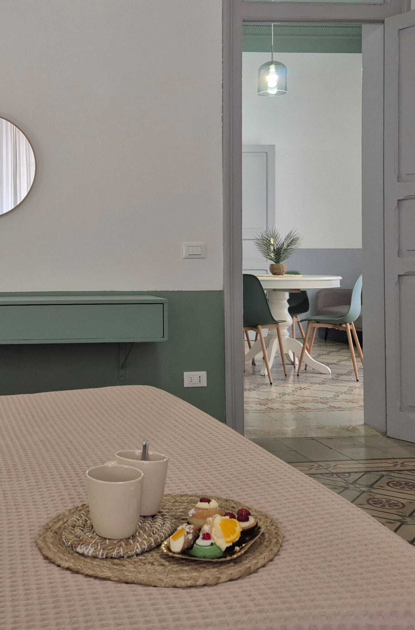 A bed with a textured bedspread, a bedside table with two white cups, and a plate of fruit.