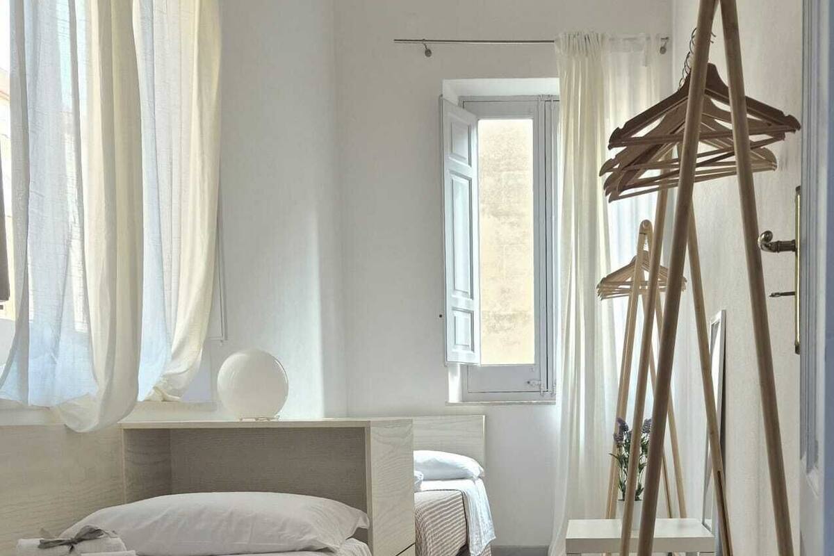 A narrow room with a bed, a window, a hanging clothes rack, and patterned floor tiles.