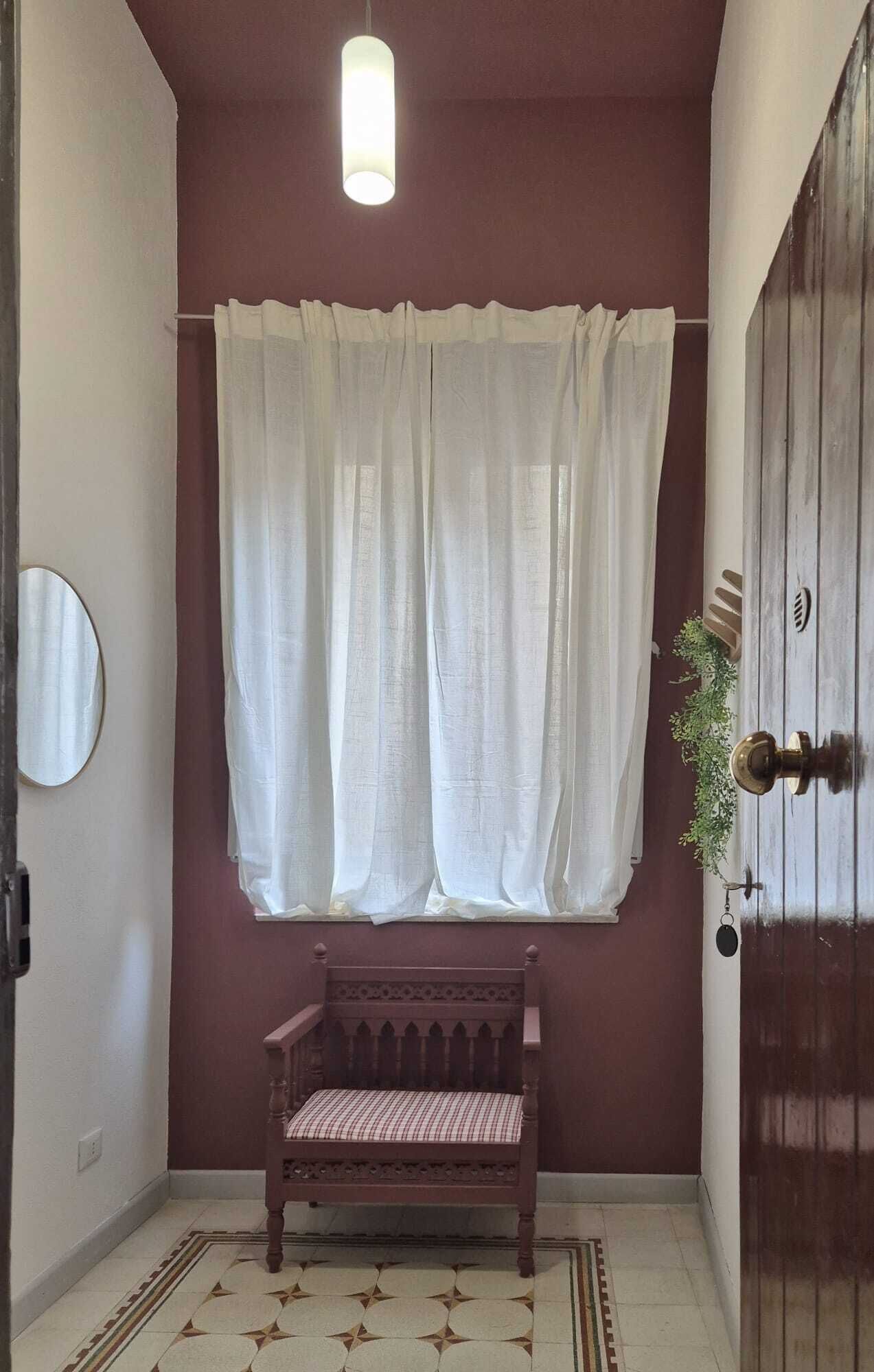 A hallway with a bench, a window with white curtains, a round mirror, and a door with a keyhole.