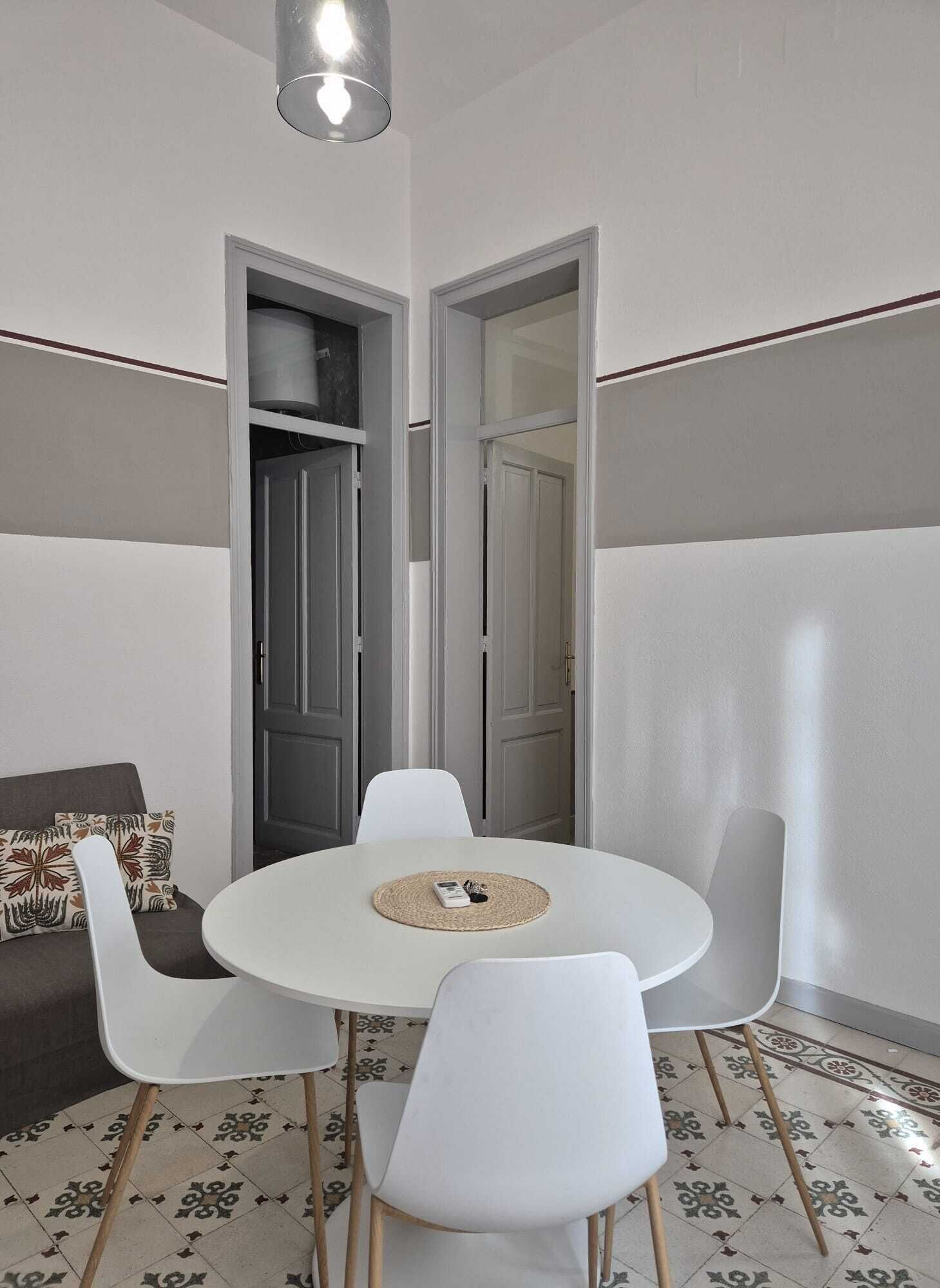 A room with a round table, white chairs, a sofa with patterned cushions, and a door leading to another room.