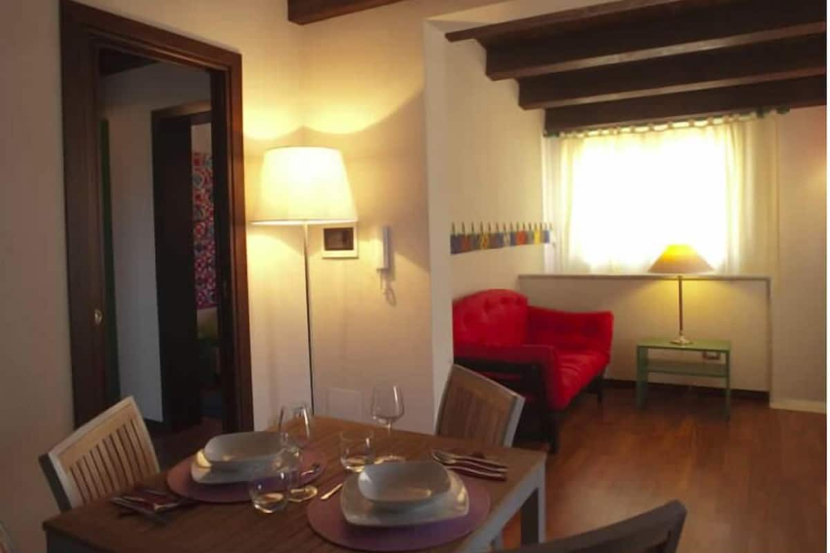 A dining area with a table set for two, a red chair, a lamp, and a window with curtains.
