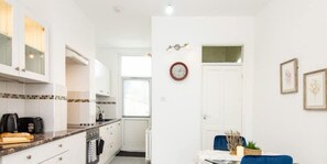 A compact kitchen with white cabinets, a coffee maker, and a toaster. There is a dining area with a table and chairs, and a clock on the wall. - Omega @ Hanwell | Ground Floor Ensuite | Parking (London)