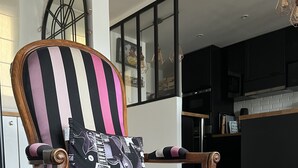 Une chaise avec un coussin à rayures et un coussin décoré d’une guitare, placés dans une pièce au plancher en bois, avec une cuisine en arrière-plan.