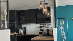 Une cuisine moderne avec des meubles noirs, un dosseret en carreaux blancs et un plan de travail en bois. On y trouve deux suspensions au design géométrique, ainsi qu’un grand présentoir noir et étroit, orné d’un motif en spirale.