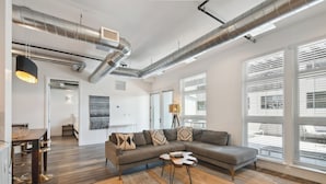 Un salon moderne avec un canapé d’angle, une table basse et un coin repas agrémenté de tabourets de bar. De grandes fenêtres équipées de stores laissent entrer la lumière, et les conduits de ventilation sont apparents au plafond.