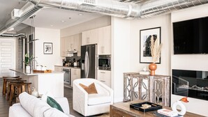 A modern living room with a sofa, a coffee table, and a TV. There is a kitchen in the background with stainless steel appliances and wooden cabinets. - Bright & Breezy | RiNo Loft with Custom Street Art | Dog-Friendly (Denver)