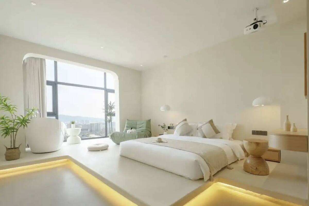 Un dormitorio moderno con un ventanal grande, una cama con ropa de cama blanca, una mesita de noche y una planta pequeña.