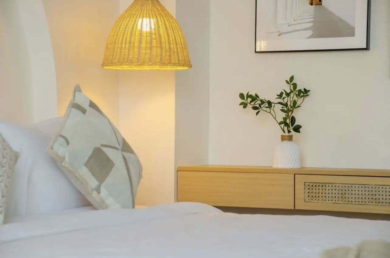 Ein Schlafzimmer mit einem Bett, einem Nachttisch mit einer Vase voller Grünpflanzen, einem Wandbild und einer Pendelleuchte.