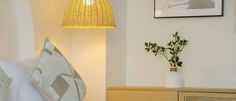 A bedroom with a bed, a bedside table with a vase of green plants, a wall-mounted picture, and a hanging lamp.