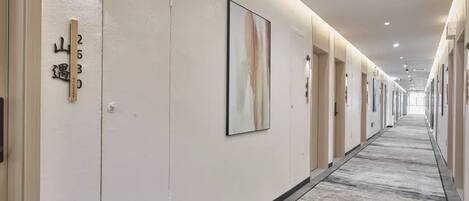 A hotel corridor with numbered doors, a framed abstract painting, and a patterned carpet.