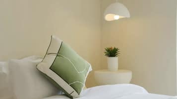 Una cama bien hecha con ropa de cama blanca y una almohada verde. Hay una pequeña planta en maceta sobre una mesa redonda blanca al lado de la cama. Una lámpara colgante blanca ilumina la cama.