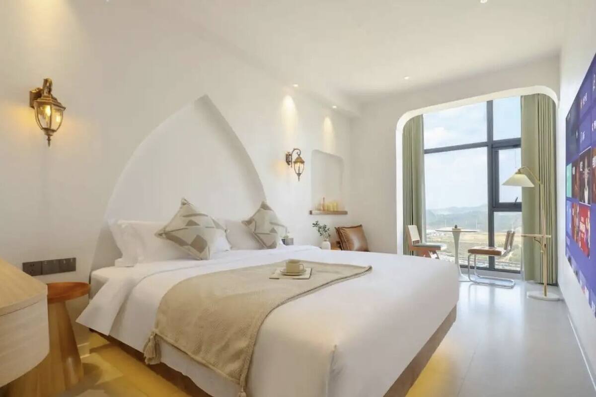 Habitación de hotel moderna con una cama grande, dos lámparas fijadas a la pared, vistas del exterior a través de un ventanal amplio y una pequeña mesa con sillas.