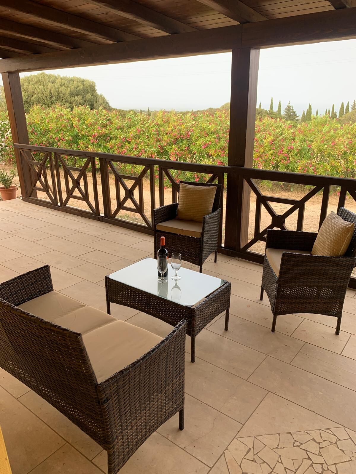 A covered patio with wicker furniture and a glass table, overlooking a garden with red flowers and trees.