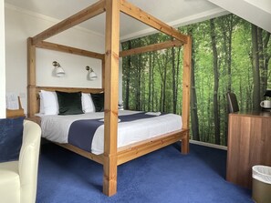 A four-poster bed with a forest wallpaper backdrop, a desk with a coffee maker, and a chair. - DERBY CITY CENTRE BUDGET HOTEL  (Derby)