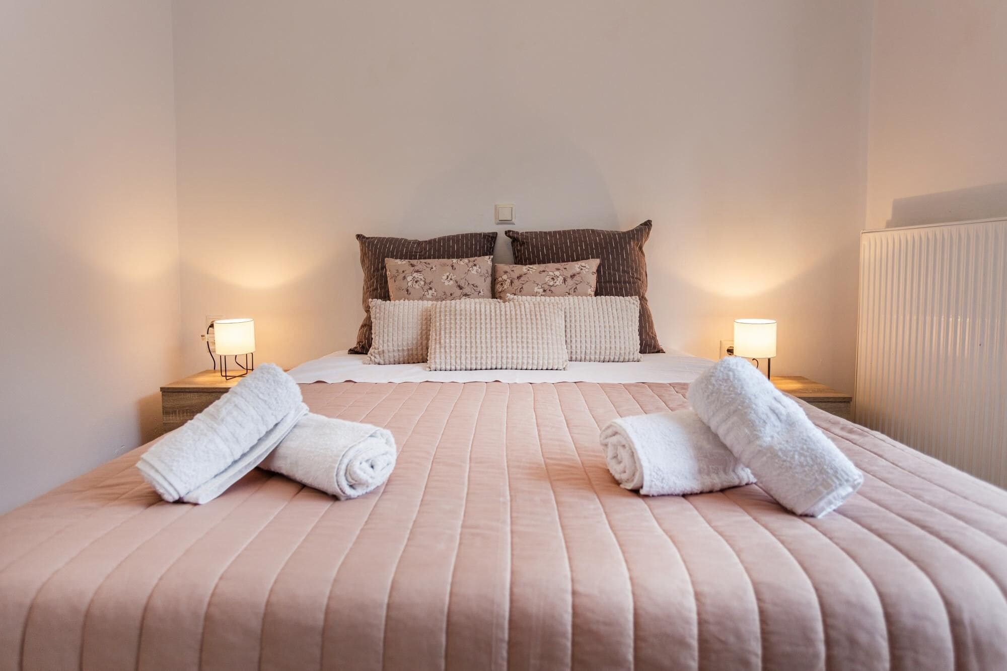 A neatly made bed with a pink bedspread and two rolled towels on the side.
