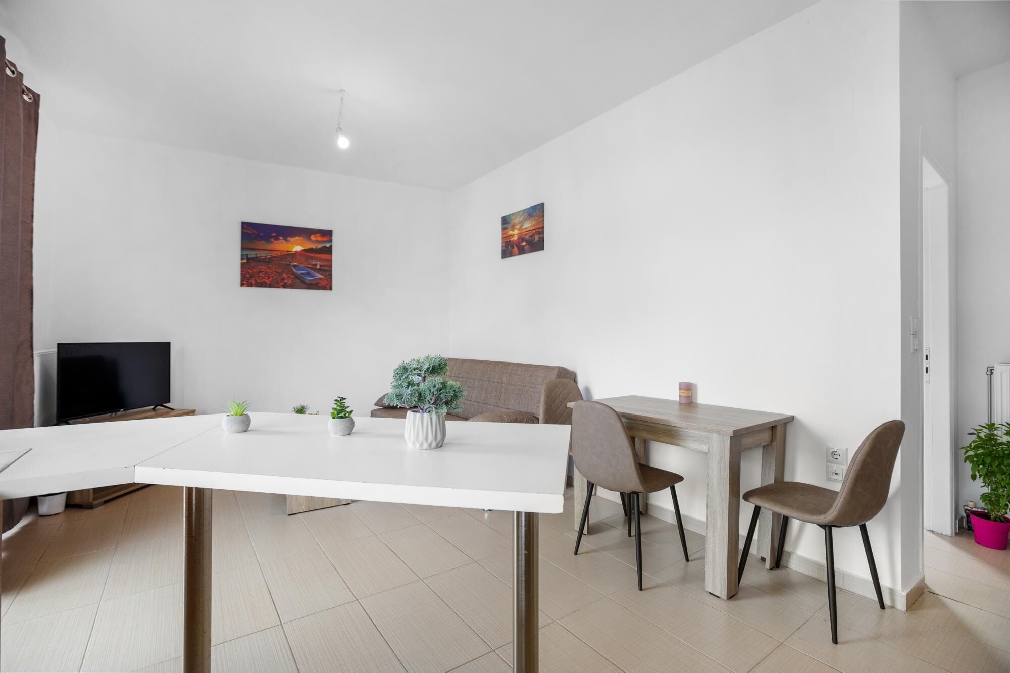 A modern living room with a white sofa, a dining table with chairs, a TV, and two paintings on the wall.