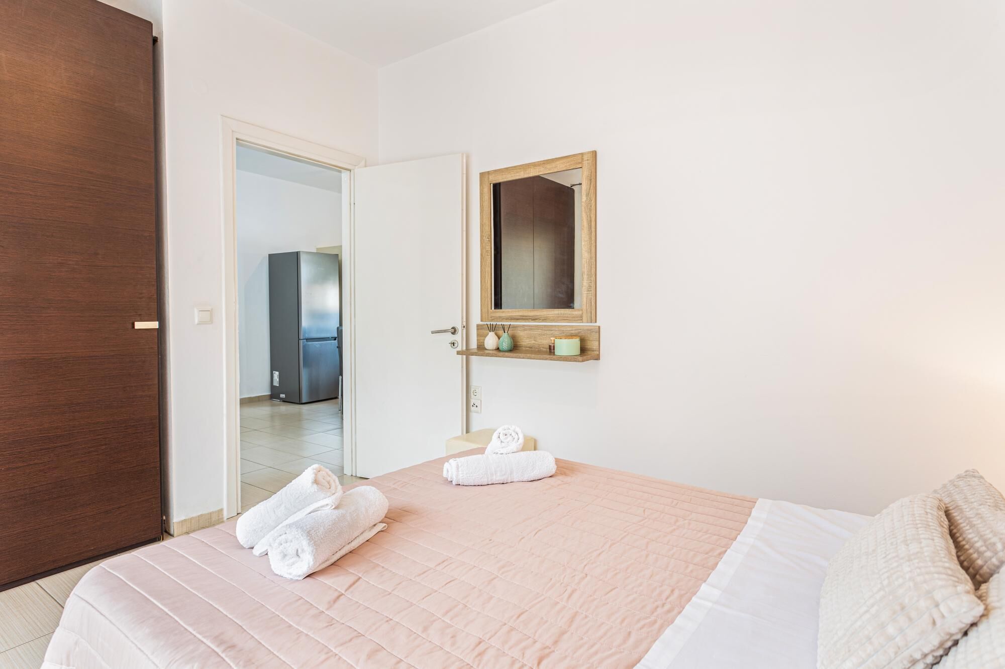 A hotel room with a bed, a wooden door, a mirror, and a small shelf with decorative items.