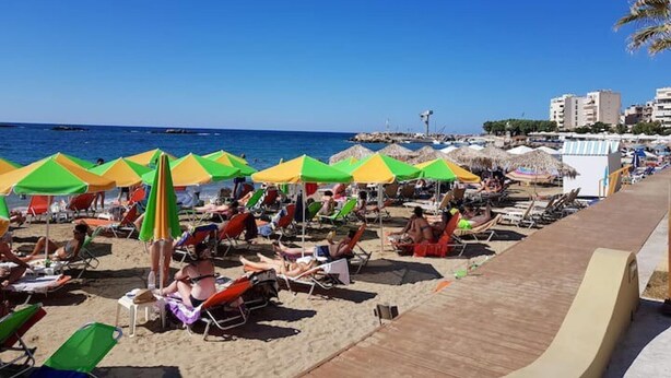 Una playa con sombrillas de colores y tumbonas, un paseo marítimo de madera y edificios al fondo.