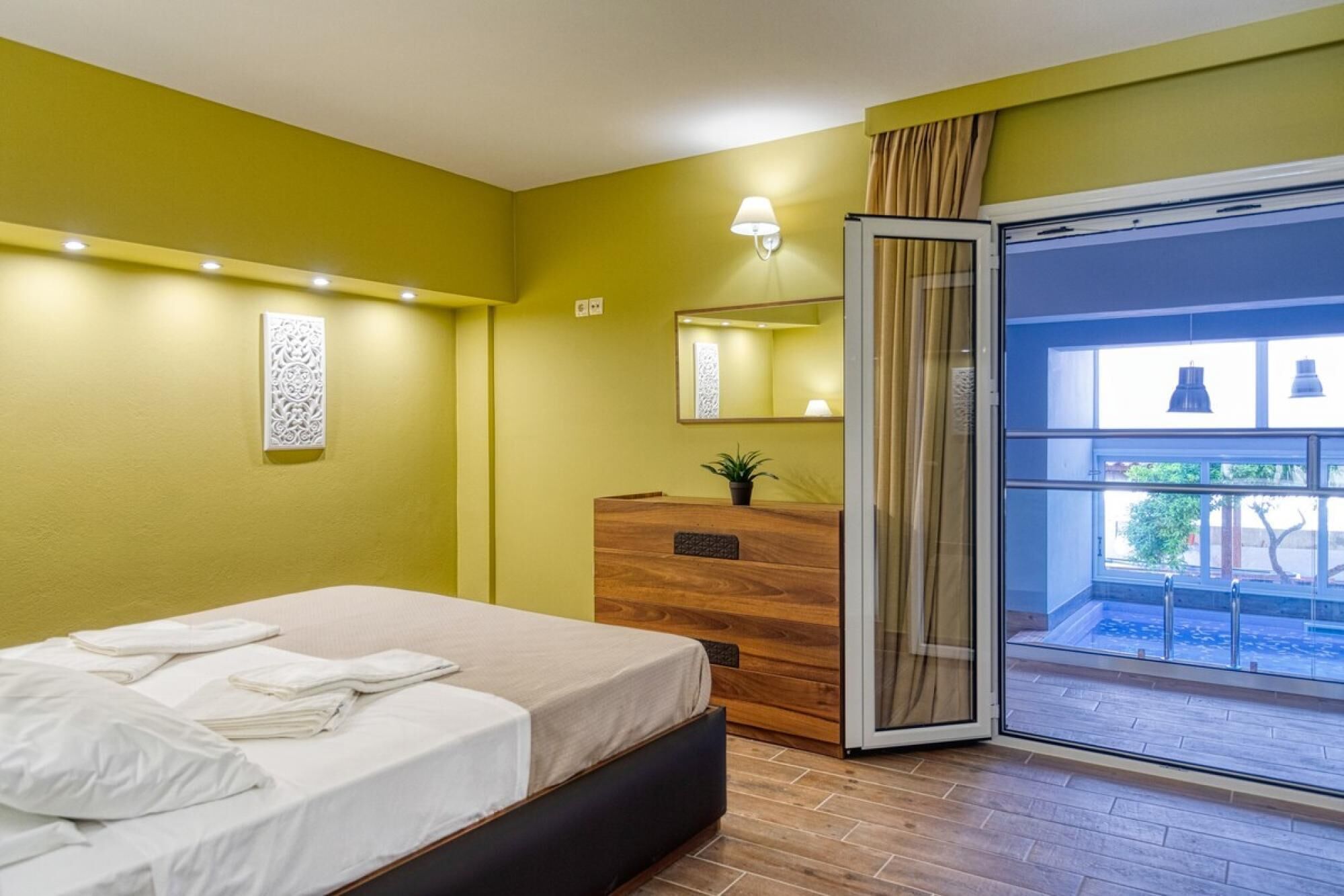 A modern hotel room with a bed, a wooden desk, a mirror, and a sliding glass door leading to a balcony.