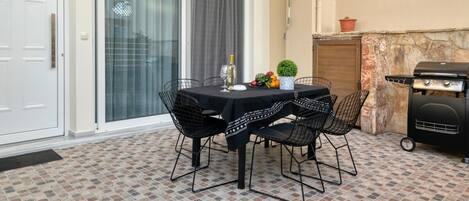 An outdoor patio with a table set for two, surrounded by chairs, a barbecue grill, and a potted plant.