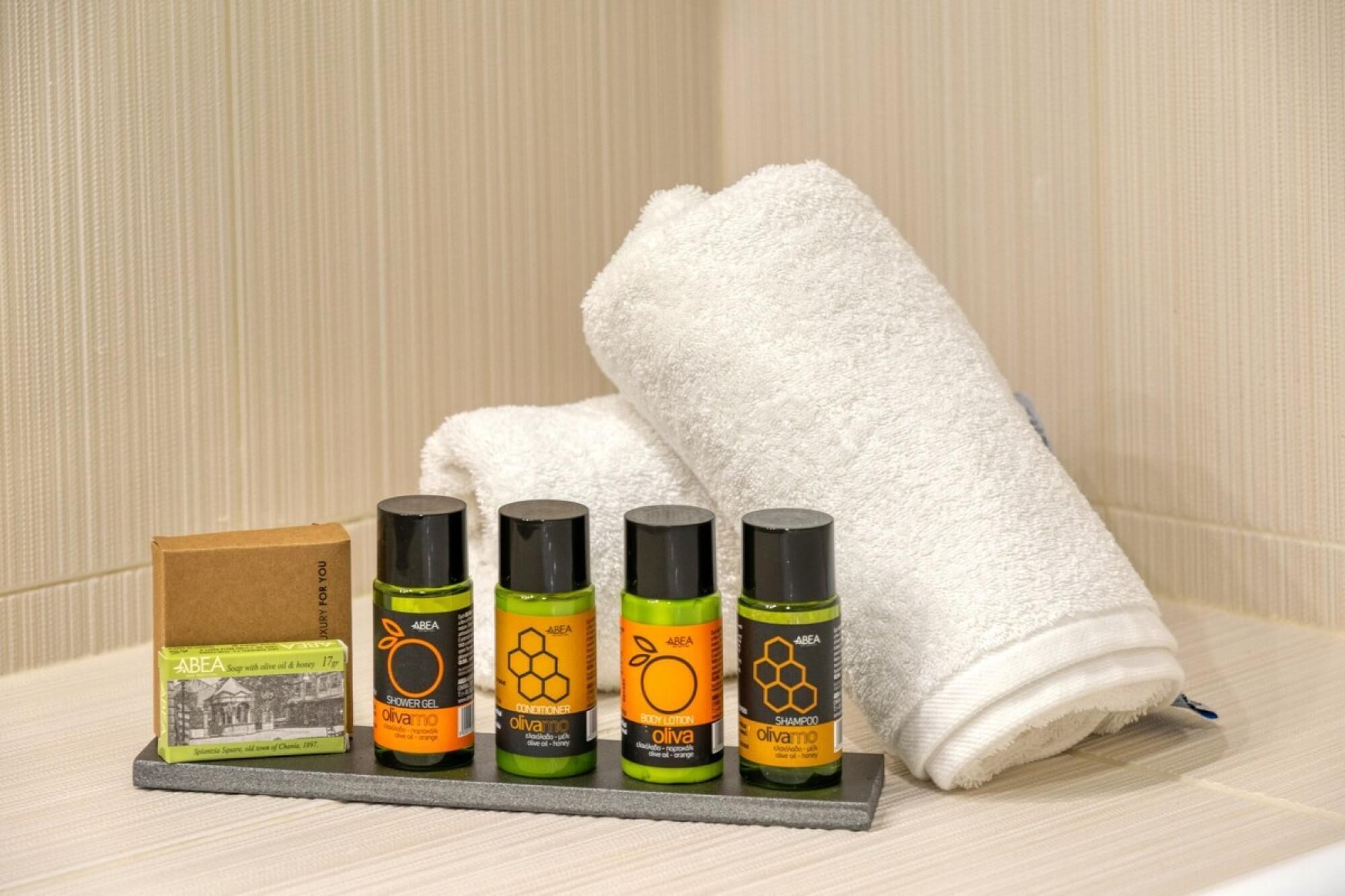 A set of toiletries including a soap bar, shower gel, conditioner, body lotion, and shampoo, arranged on a tray with a white towel in the background.