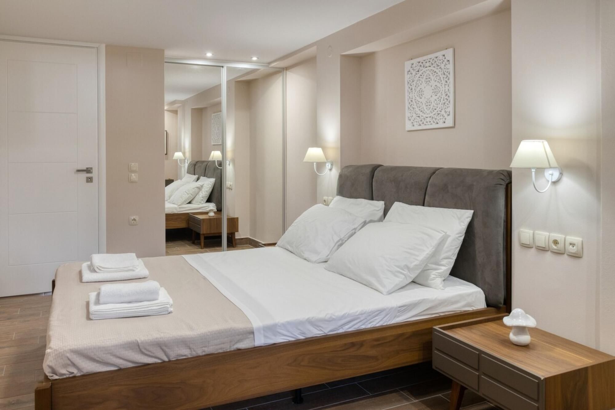 A modern hotel room with a wooden bed, white linens, a nightstand with a lamp, and a mirror reflecting the room.