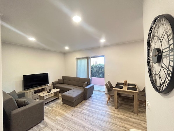 A modern living room with a flat-screen TV, a sofa, a coffee table, and a dining area with a clock on the wall. - Venetian Kamara in Old Town Chania (Chania)