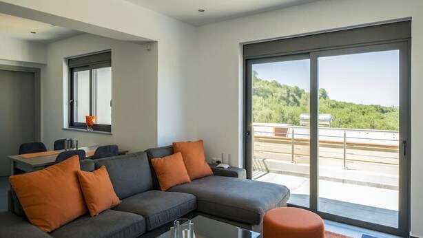 A modern living room with a grey sofa, orange cushions, a glass coffee table, and a large sliding glass door leading to an outdoor balcony with a view of greenery. - Kayzo Luxury Apartment With Pool (Platanias)