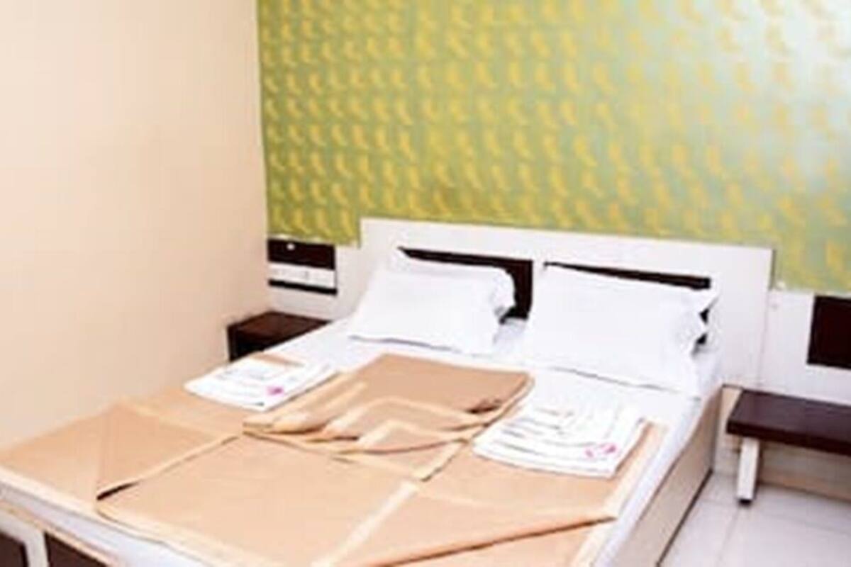A neatly made bed with a white headboard, a brown blanket, and a white pillow. There is a small bedside table with a lamp and a wall-mounted air conditioner.