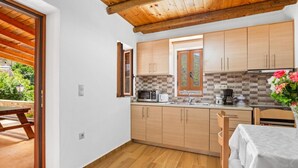 A compact kitchen with wooden cabinets, a tiled backsplash, and a window with a view of greenery. - Maria s House With View in Kambos (Kissamos)