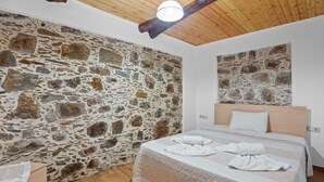 A bedroom with a stone wall, wooden ceiling beams, a bed with white linens, and a wooden dresser. - Maria s House With View in Kambos (Kissamos)