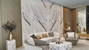 A modern living room with a marble accent wall, a grey sofa, a matching armchair, and a marble coffee table. - Luxe 1BR 15Mins- Beach Best Infinity Pool -Jumeirah Village Circle ,JVC  (Dubai)