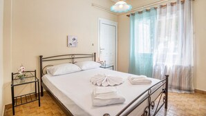 A neatly made bed with white linens and towels, a bedside table with a small vase, a wall-mounted picture, and a window with sheer curtains. - Ortansia House Near the Sea (Platanias)