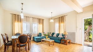 A spacious living room with a dining area, large windows, and a view of greenery outside. - Ortansia House Near the Sea (Platanias)