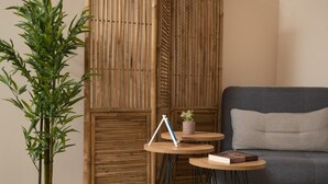 A room with a bamboo partition, a grey sofa, and two small wooden tables with books and a plant. - Harmony Apartment in Chania (Chania)