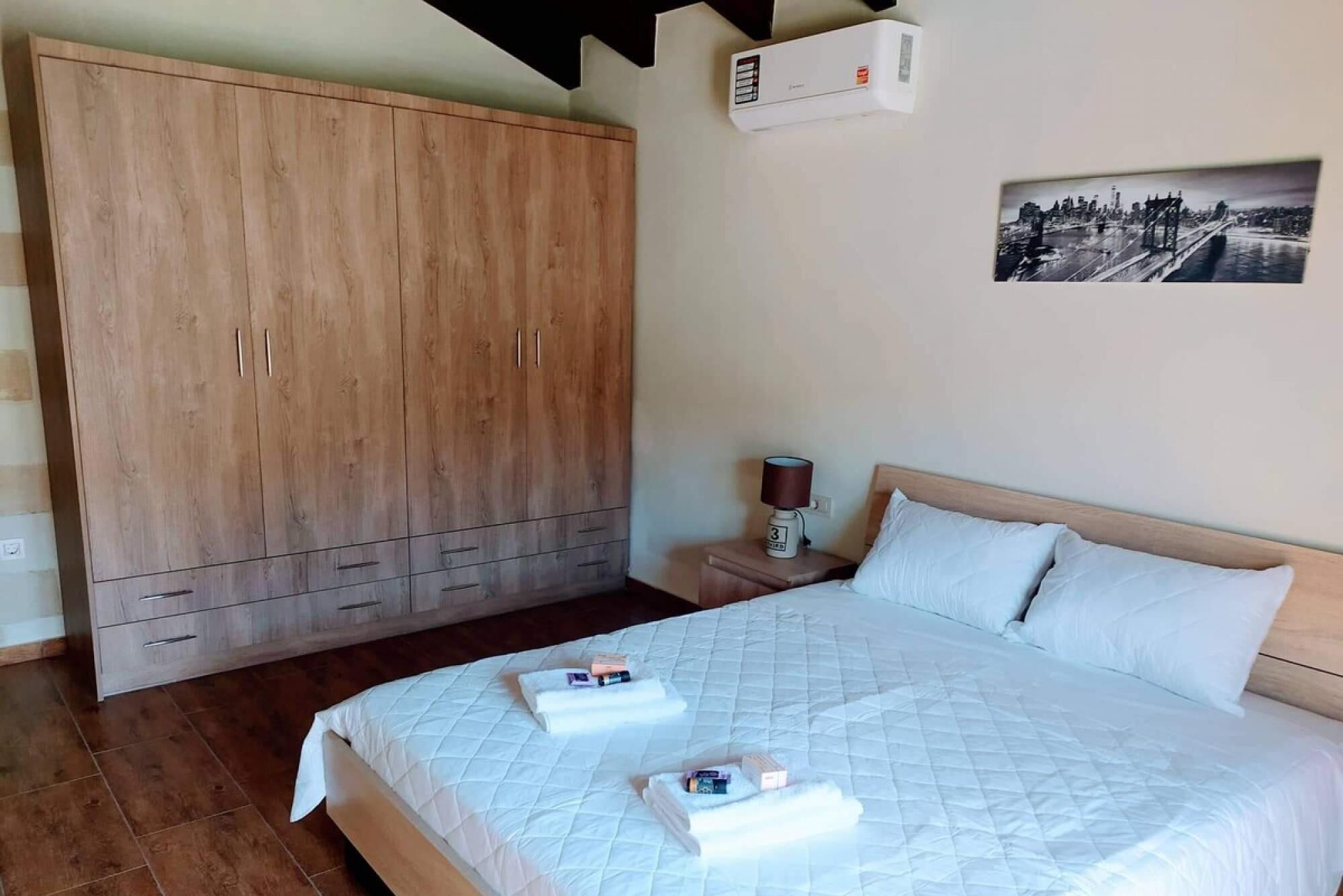 Een slaapkamer met een grote houten garderobe, een bed met wit beddengoed, een nachtkastje met een lamp en een foto van een stadsgezicht aan de muur.