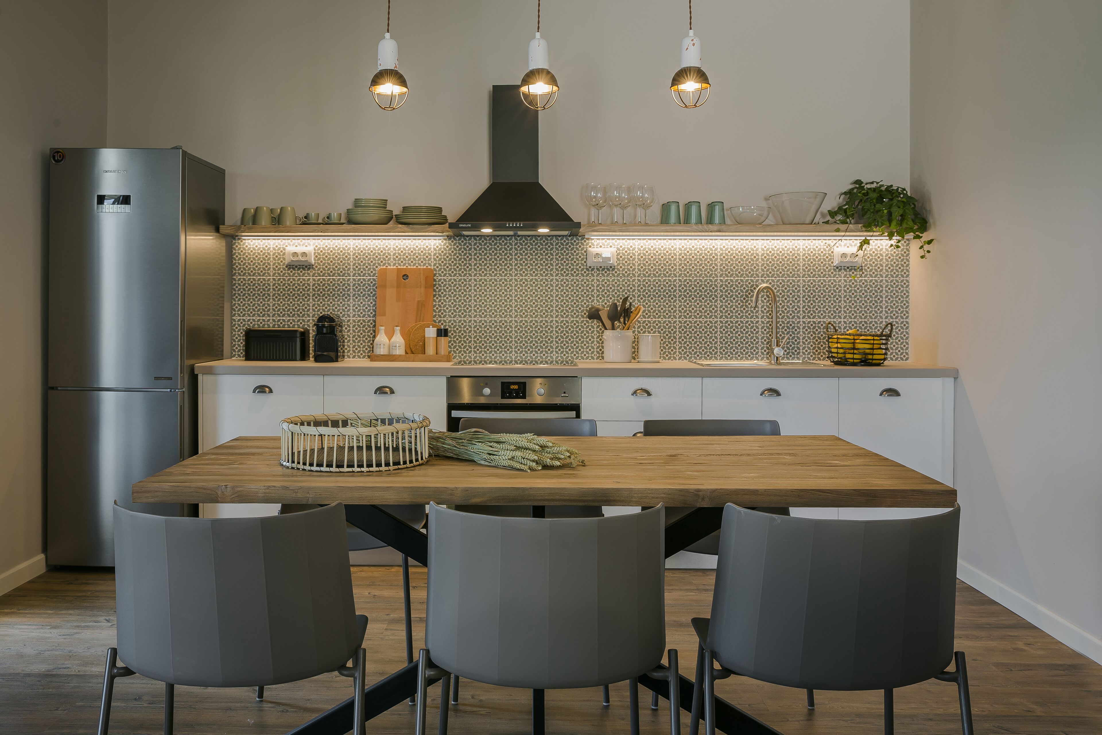 Una cocina moderna con una mesa de madera, sillas grises, un refrigerador de acero inoxidable y un salpicadero con diseño.