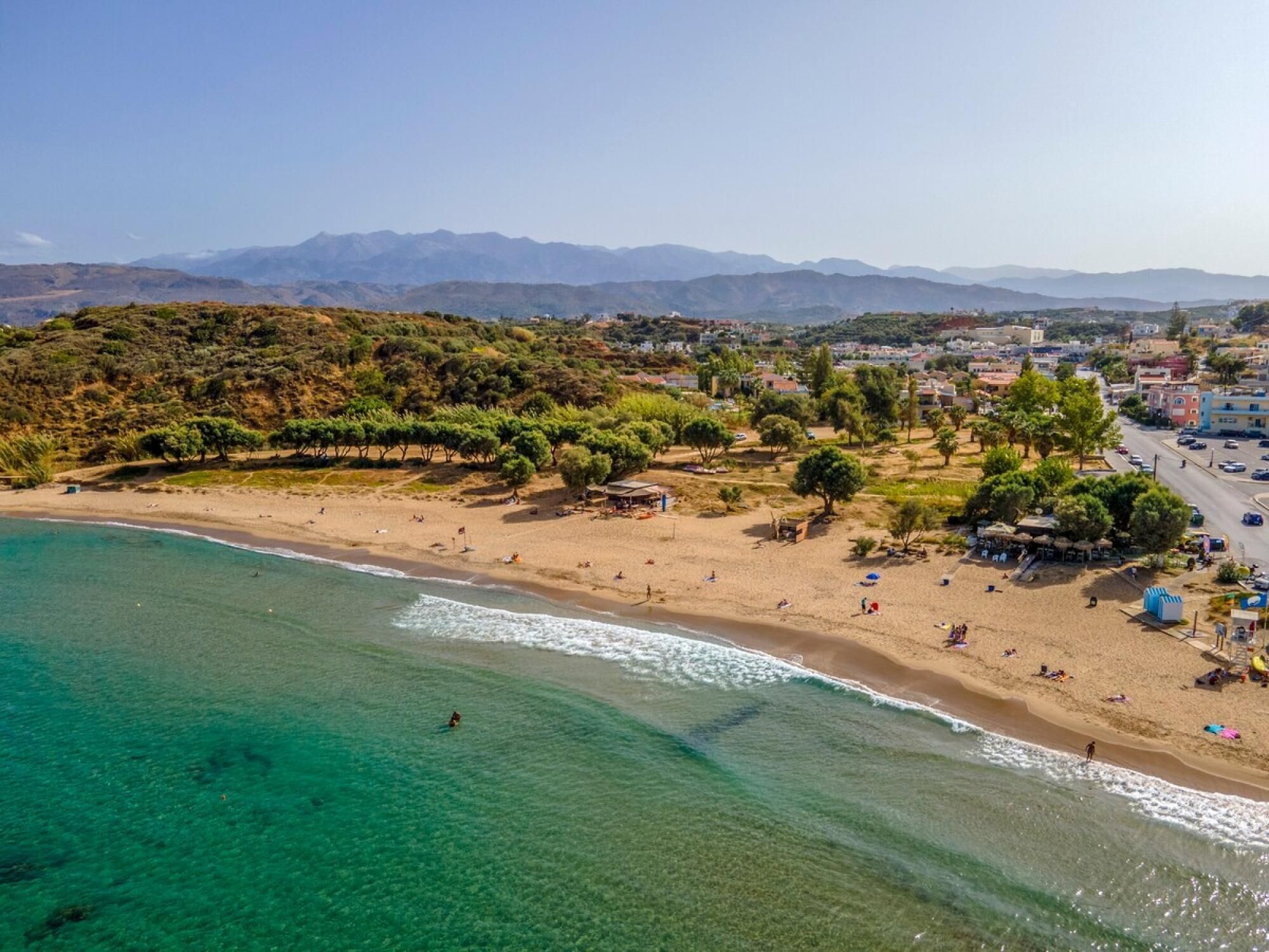 A coastal beach with clear turquoise waters, a sandy shore, and a backdrop of hills and buildings.