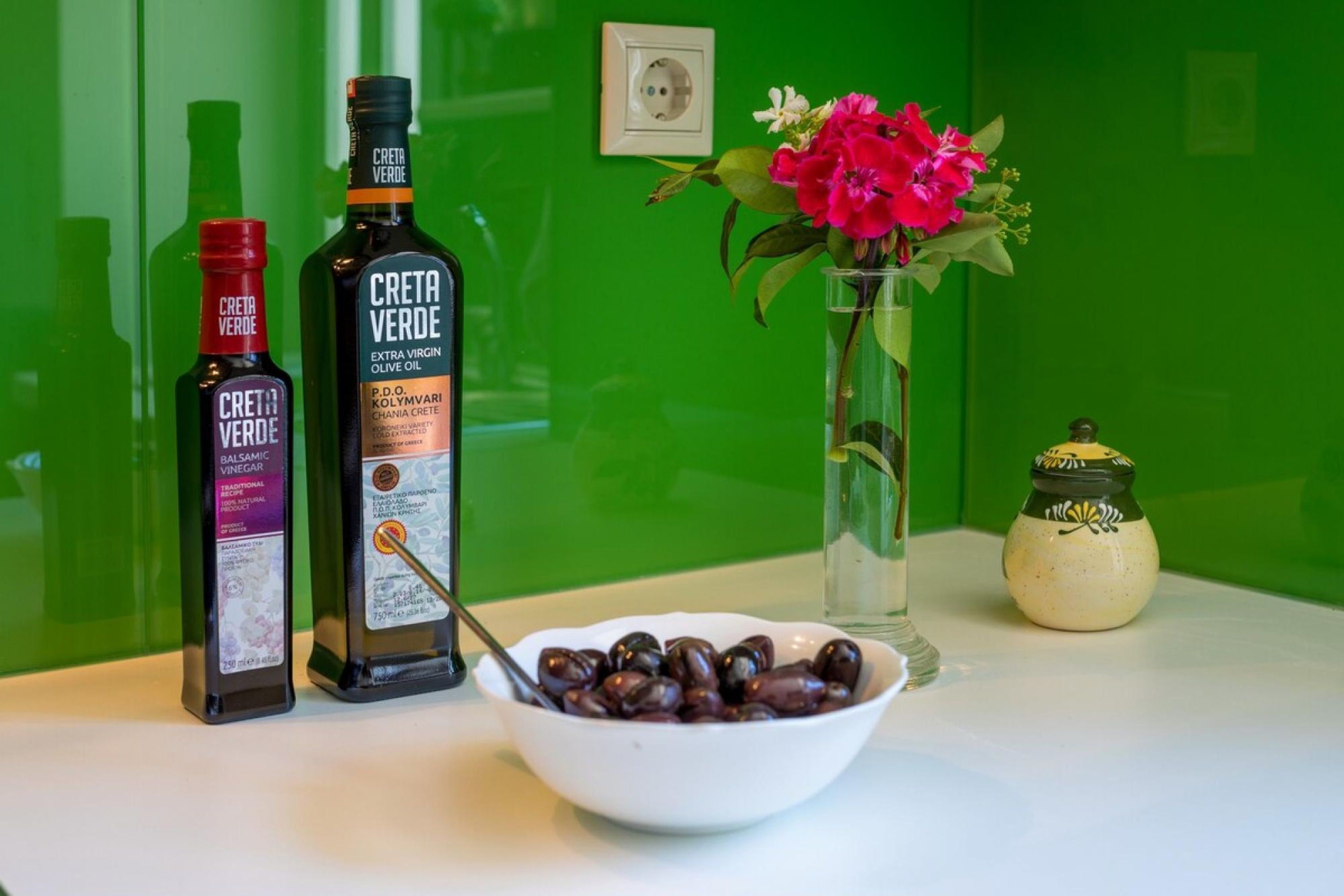 Dos botellas de aceite de oliva virgen extra Creta Verde, un cuenco de aceitunas, un jarrón con flores y un frasco pequeño sobre una mesa frente a un muro verde.