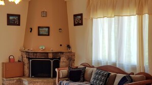 Ein gemütliches Wohnzimmer mit Kamin, einer Couch mit gemusterten Kissen und einem Fenster mit durchsichtigen Vorhängen.
